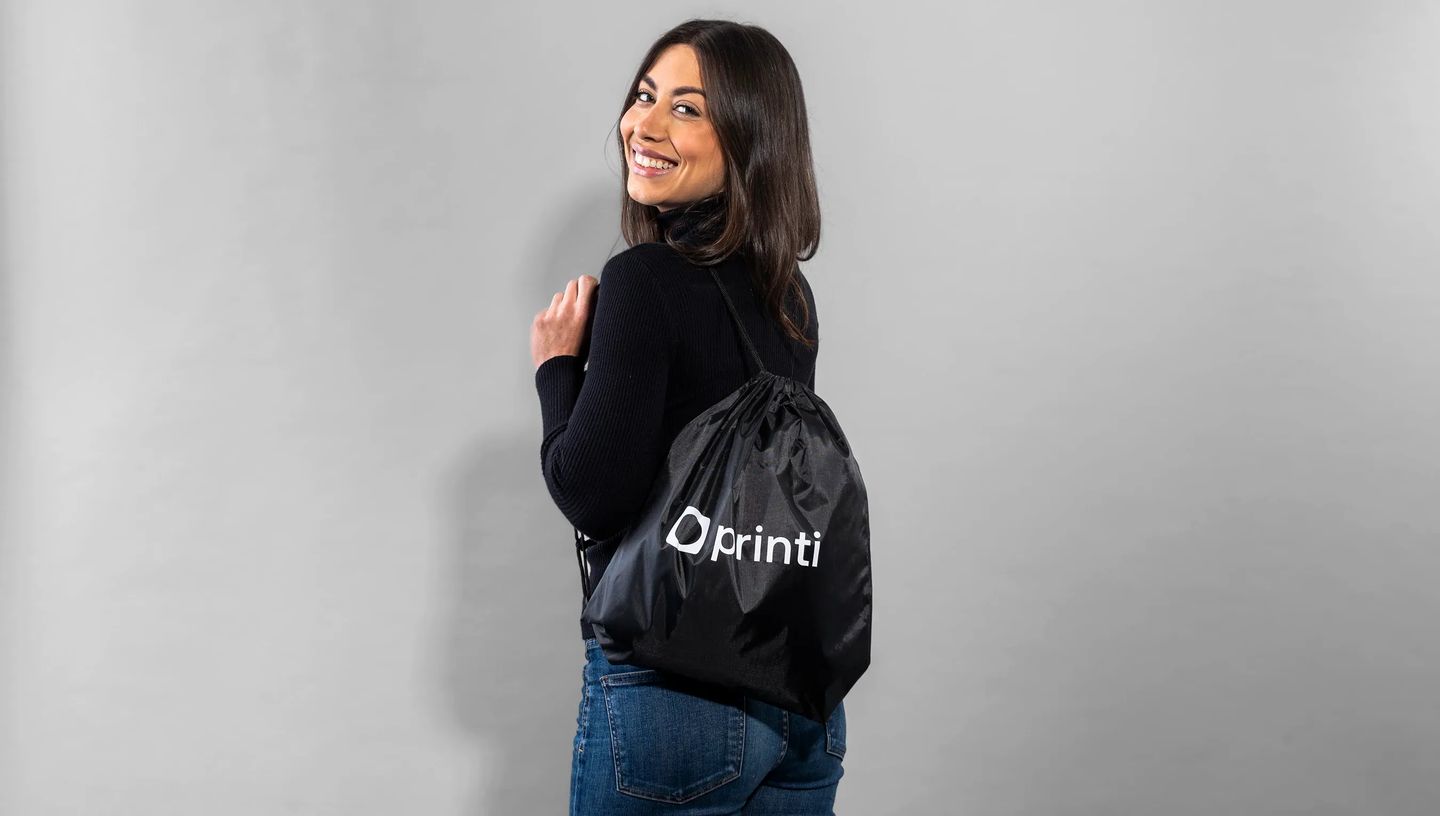 mulher branca, usando uma calça jeans e casaco preto, posicionada olhando para trás e sorrindo, euquando usa uma mochila saco personalizada na cor preta com logo da Printi