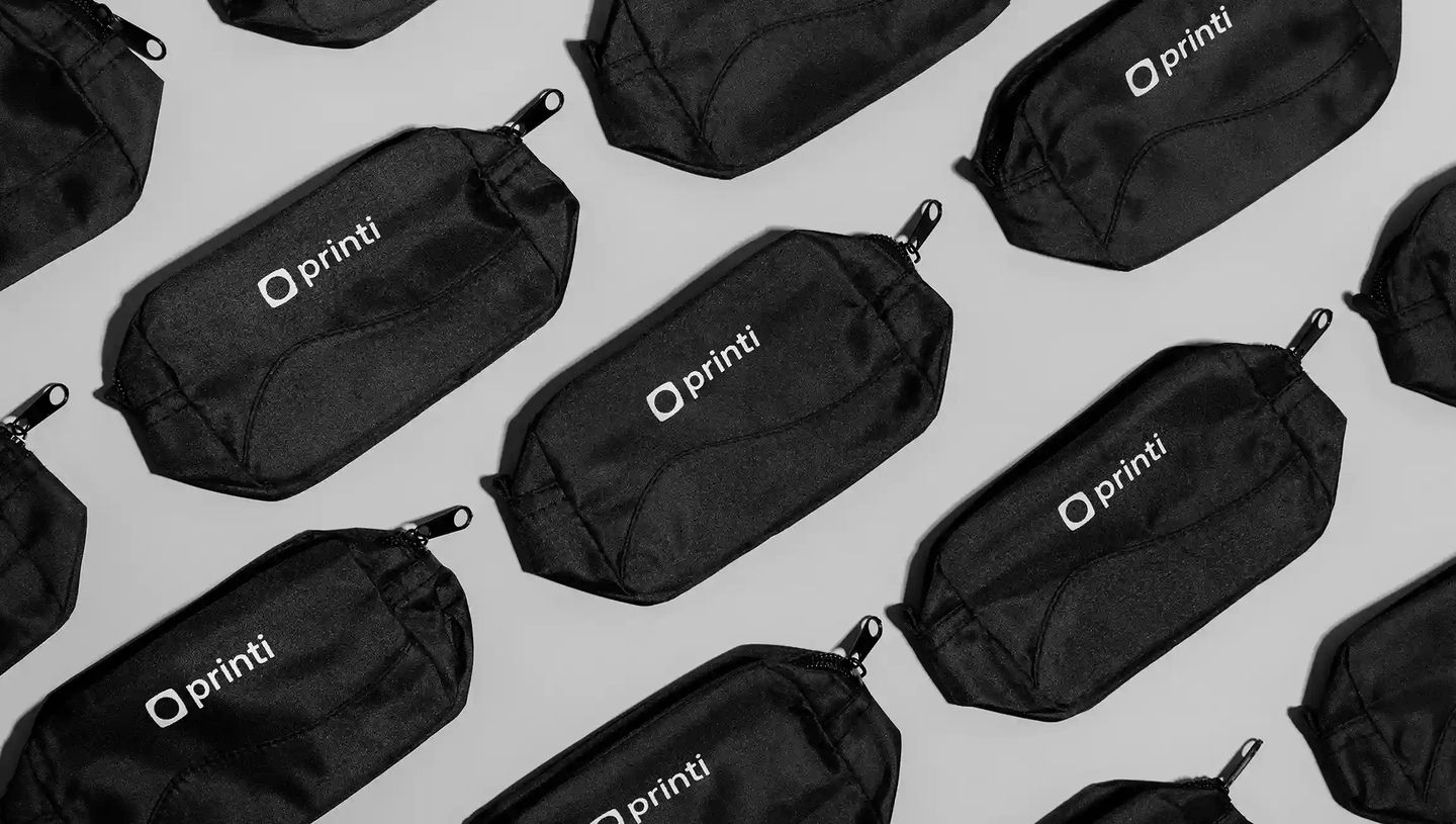 Necessaire personalizada preta com zíper e logo Printi em destaque, exibida em padrão repetido sobre fundo neutro, destacando o design funcional do estojo organizador para itens pessoais ou corporativos.