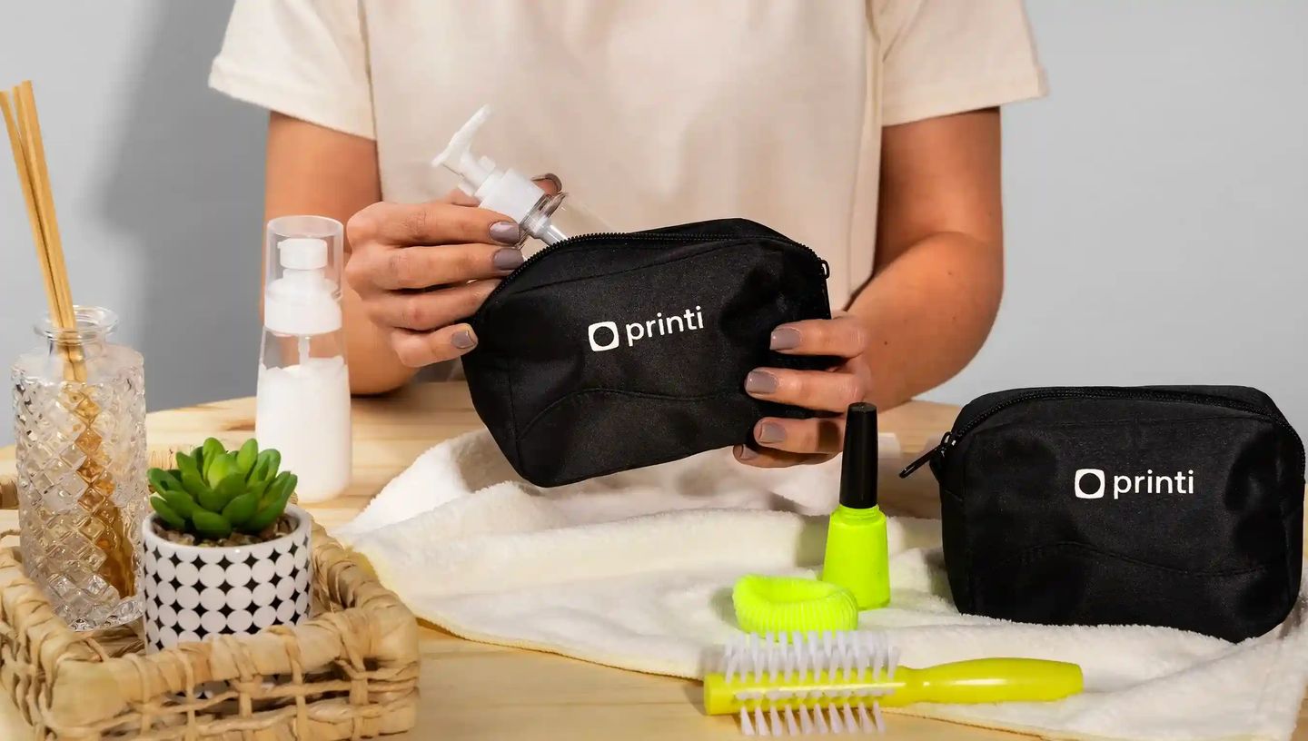 Necessaire personalizada preta com logo Printi sendo aberta para guardar frasco de sabonete e itens de cuidados pessoais.