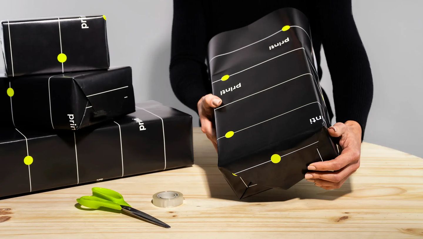 Pessoa embrulhando caixas de presente com papel de presente preto personalizado com detalhes gráficos em linhas brancas e pontos verde neon da Printi. As caixas aparecem sobre uma mesa de madeira junto com tesoura e fita adesiva