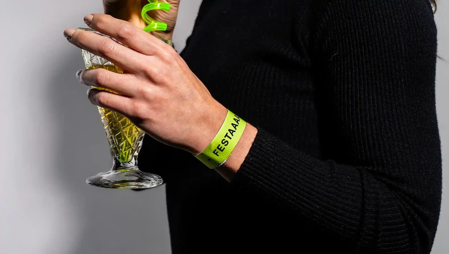 Pulseira de identificação personalizada verde sendo usada no pulso em evento, enquanto a pessoa segura um copo de bebida.