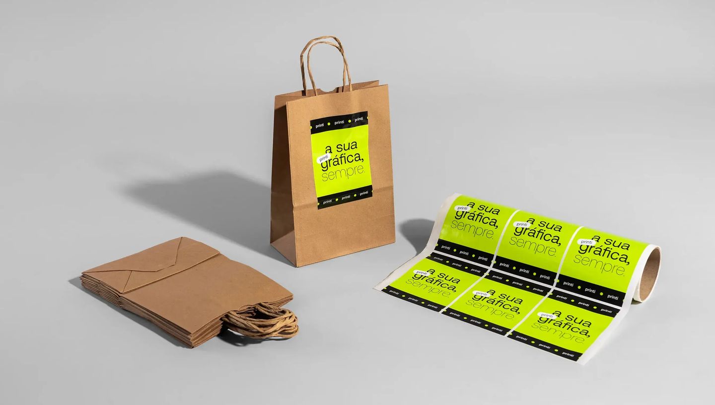 Sacola de papel kraft personalizada com adesivo verde neon da Printi aplicada na frente, acompanhada de pilha de sacolas e rolo de adesivos personalizados