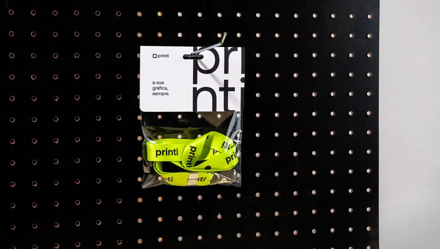 Solapa personalizada da Printi presa em painel perfurado exibindo cordão promocional verde-limão com logo Printi dentro de embalagem transparente, utilizada para apresentação de brindes, identificação de produtos e materiais promocionais em lojas e evento