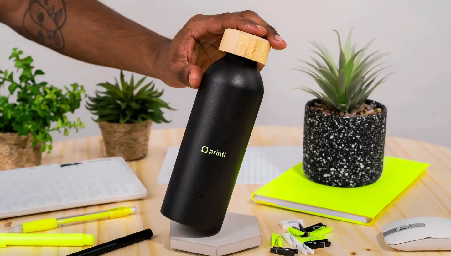 Squeeze térmico preto personalizado com tampa de bambu sendo segurado sobre mesa de escritório com caderno, canetas e plantas.