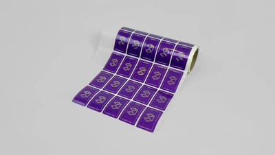 Rolo de adesivos personalizados quadrados com fundo roxo e impressão branca, parcialmente desenrolado sobre fundo neutro, mostrando várias etiquetas prontas para aplicação em embalagens ou produtos.