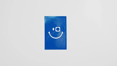 Adesivo retangular personalizado em fundo azul com acabamento brilhante e ilustração gráfica branca em formato de rosto sorridente, utilizado para rotulagem de produtos, embalagens, brindes e comunicação visual de marcas.