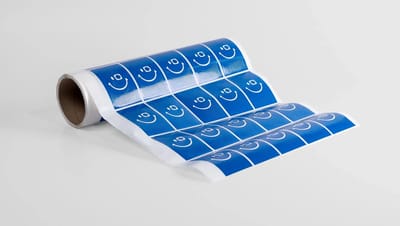 Rolo parcialmente desenrolado de adesivos retangulares azuis organizados em cartela, cada um com ícone de rosto sorridente em branco, exibido sobre fundo neutro e representando etiquetas adesivas personalizáveis ideais para embalagens, identificação de pr