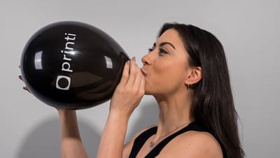 Mulher branca de cabelo preto liso, posicionada de lado, inflando balão personalizado preto com logotipo da Printi