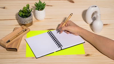 Pessoa escrevendo em um caderno espiral com uma caneta do Conjunto de Caneta e Lapiseira em Bambu, posicionada em um ambiente de trabalho com decoração minimalista,
