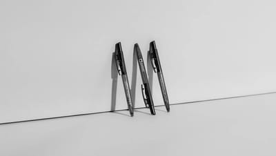 Três canetas plásticas transparentes personalizadas com logotipo da Printi e detalhes em preto, apoiadas sobre superfície neutra com iluminação suave, representando brindes corporativos e materiais promocionais ideais para empresas, eventos, ações de mark