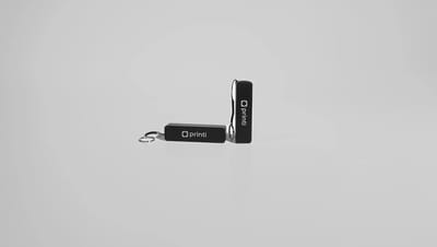 Dois carregadores portáteis personalizados com logo Printi posicionados sobre fundo neutro, um na horizontal com chaveiro metálico e outro em pé com cordão de transporte, representando um brinde tecnológico compacto e funcional para recarregar smartphones