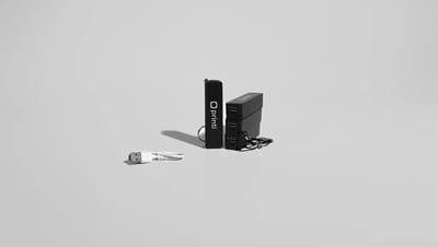 Carregadores portáteis personalizados da Printi em cor preta com logotipo branco, acompanhados de cabo USB, exibidos sobre fundo neutro como exemplo de brinde corporativo tecnológico ideal para eventos, ações promocionais, kits de onboarding e estratégias