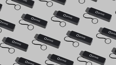 Carregadores portáteis personalizados da Printi na cor preta, com logo em branco e cordão com argola metálica para chaveiro, repetidos em padrão sobre fundo neutro, representando power banks promocionais usados como brindes corporativos para recarregar sm