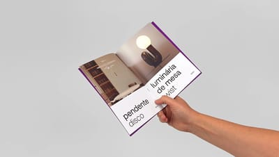Catálogo personalizado impresso sendo segurado por uma mão, aberto em página com fotos de luminária e design editorial moderno.