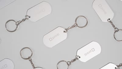 Diversos Chaveiros de Metal Básico com plaquinha retangular prateada personalizada com logo da Printi, exibido em padrão repetido sobre fundo cinza claro