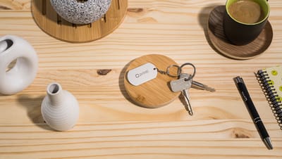 Chaveiro Metal Básico com plaquinha de alumínio personalizada com logo da Printi, apoiado sobre porta-chave de bambu em mesa de madeira clara com objetos decorativos ao redor
