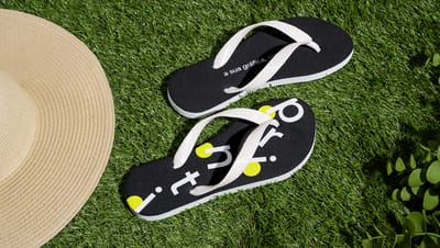 Chinelo personalizado na cor preta com alça branca perto de chapéu em grama sintética verde.