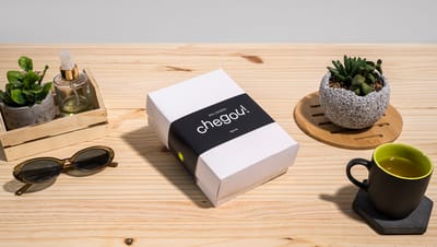 Cinta para embalagem tampa e fundo na cor preta com a frase “seu pedido chegou!”, aplicada em uma caixa branca sobre uma mesa de madeira com objetos decorativos