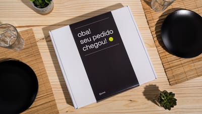 Caixa de transporte branca com cinta preta em mesa de madeira perto de copo, vaso de planta e pratos pretos