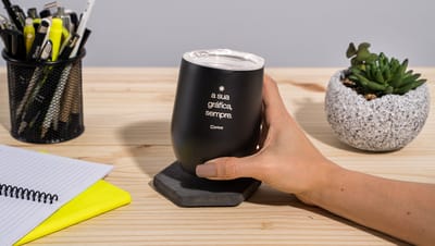 Mão de uma pessoa branca apoiada em uma superfície de madeira segurando um Copo Térmico de 400 ml preto com tampa, personalizado com a frase "a sua gráfica, sempre", com um papel branco e amarelo ao lado, um porta lápis com diversas canetas e lápis dentro