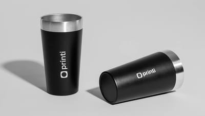 Dois copos térmicos personalizados com a logo da Printi, na cor preta e sem tampa, com acabamento em inox, um em pé e outro deitado ao lado