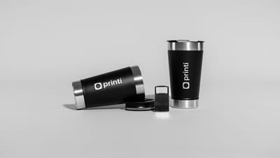 Dois copos térmicos com tampa personalizados da marca Printi, sendo um copo em pé e um deitado, com acabamento preto fosco e detalhes metálicos em aço inox