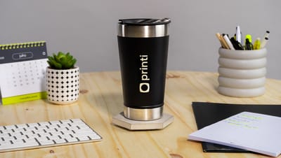 Copo térmico com tampa sobre uma mesa de madeira de escritório, ao lado de um teclado, bloco de notas, calendário, porta lápis e um vasinho branco com planta