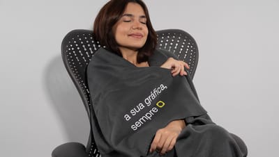Mulher sentada em cadeira utilizando a manta polar preta com slogan da Printi