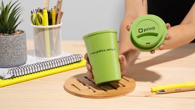 Pessoa segurando copo em fibra de bambu e PP verde da marca Printi sobre mesa de madeira com itens de papelaria ao fundo.