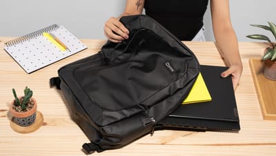 Pessoa colocando materiais dentro da mochila para notebook Birmingham 15.6 em mesa de madeira com calendário e planta ao redor.