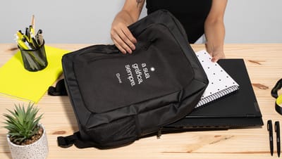 Pessoa colocando um caderno dentro de uma mochila preta sobre uma mesa com itens de papelaria e uma suculenta.