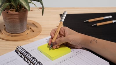 Pessoa com caneta em bambu beal na mão escrevendo em bloco de notas, em cima de caderno sobre mesa de madeira com vaso de planta e duas canetas perto..