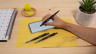 Pessoa utilizando uma caneta touch em um smartphone sobre uma mesa de madeira com bloco de notas, outras canetas, planta decorativa e itens de papelaria.