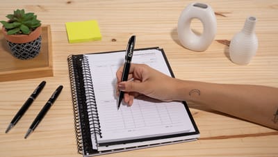 Pessoa escrevendo em caderno com caneta de metal lando personalizada na cor preta sobre mesa de madeira.