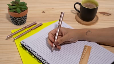 Pessoa escrevendo em caderno com caneta de metal clare sobre mesa de madeira.