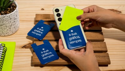 Pessoa segurando celular com porta cartão na cor azul e cartão na cor verde, em mesa de madeira com outros porta cartões.
