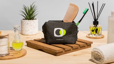 Necessaire preta com estampa personalizada apoiada em base de madeira, cercada por itens de cuidados pessoais como escova, toalha, óleos aromáticos, difusor e planta decorativa.