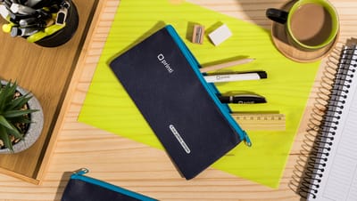 Estojo simples nas cores azul claro e escuro, com material escolar ao redor e xícara de café.