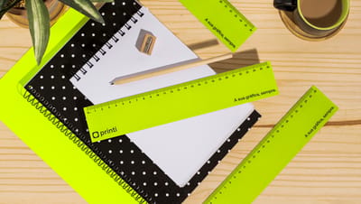 Réguas de papel na cor verde com logo da Printi junto com material escolar e de papelaria em mesa de madeira.