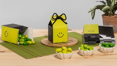 Embalagem milk com smile, na cor verde e preto ao lado de outras embalagens milk em mesa, com doces e planta ao redor