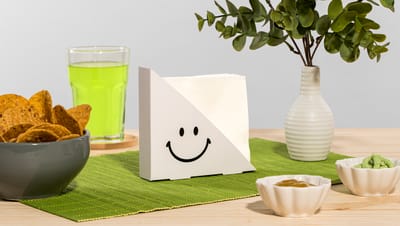 Porta-guardanapo branco com estampa de smiley, apoiado sobre jogo americano verde, ao lado de tigelas com petiscos, bebida verde e vaso decorativo.