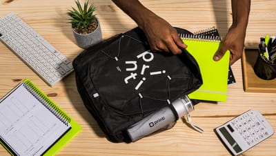 Pessoa colocando caderno dentro de mochila básica com garrafa, com caderno, teclado, suculenta, calculadora e kit de escrita ao redor.