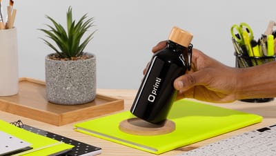Pessoa posicionando um squeeze preto de alumínio reciclado com tampa de madeira e logo da Printi sobre um caderno em uma mesa de escritório.