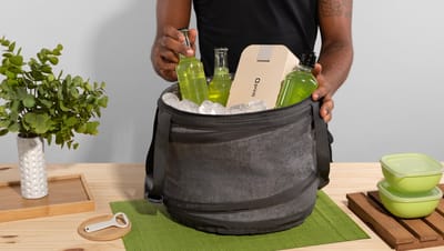 Pessoa colocando uma garrafa de bebida dentro de uma bolsa térmica cinza cheia de gelo em mesa de madeira com planta e coisas de cozinha ao redor.
