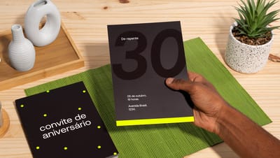Pessoa segurando um convite de aniversário preto com o número 30 em destaque, sobre uma mesa com outro convite, suculenta e objetos decorativos.