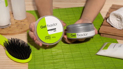 Pote de gel com rótulo personalizado em toalha verde com escova e produtos para cabelo sobre mesa de madeira.
