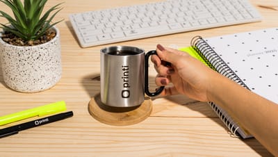 Pessoa segurando caneca para transporte sobre mesa com planta e cadernos ao redor.