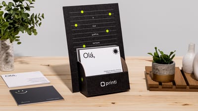 Display take one personalizado na cor preta com cartão de visita dentro e com plantas ao redor sobre mesa de madeira.