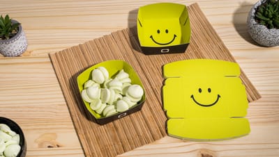 A imagem mostra embalagens bandeja de papel cartonado, com design amarelo e preto e estampa de smile. Duas estão montadas (uma com doces) e uma aberta, exibindo o molde. Elas estão dispostas sobre uma esteira de bambu com fundo de madeira clara, ao lado d