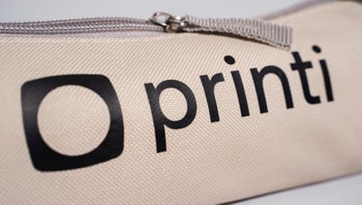 Detalhe do estojo para lápis com tecido resistente em tom bege, zíper metálico e logotipo da Printi estampado em preto